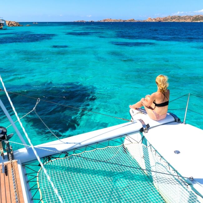 Woman in bikini tanning and relaxing on a summer sailin cruise, sitting on a luxury catamaran in picture perfect turquoise blue lagoon near Spargi island in Maddalena Archipelago, Sardinia, Italy.